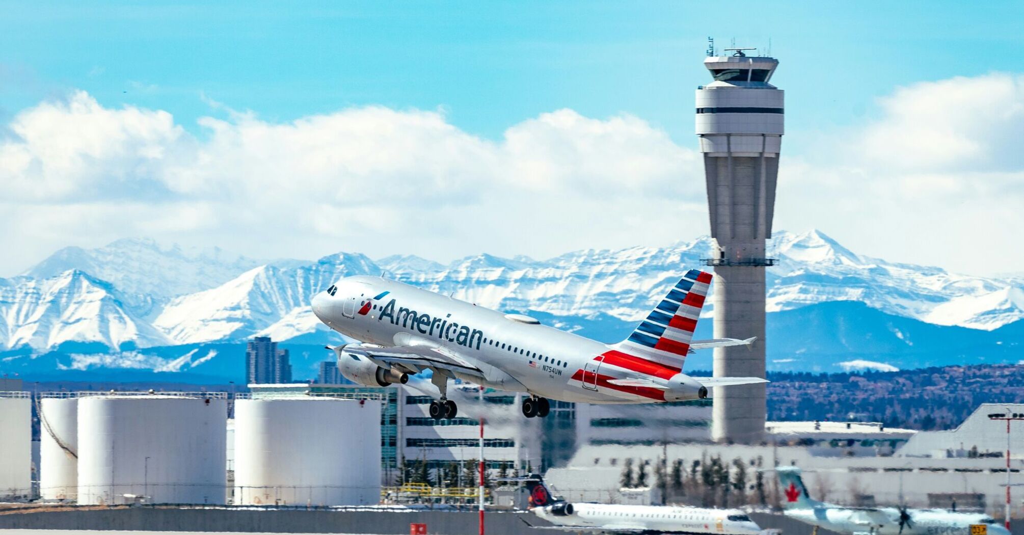 American Airlines aircraft taking off from an airport with a control tower in the foreground and snow-capped mountains in the background