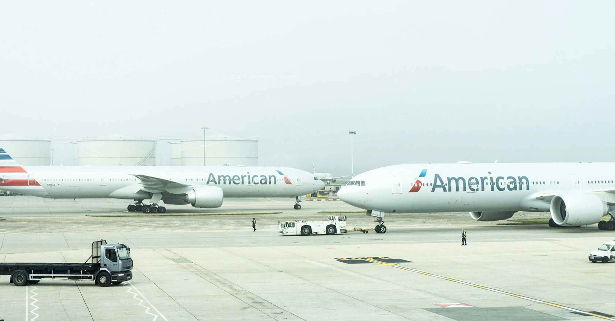 Two American Airlines aircraft parked at an airport on a hazy day with ground vehicles in the foreground