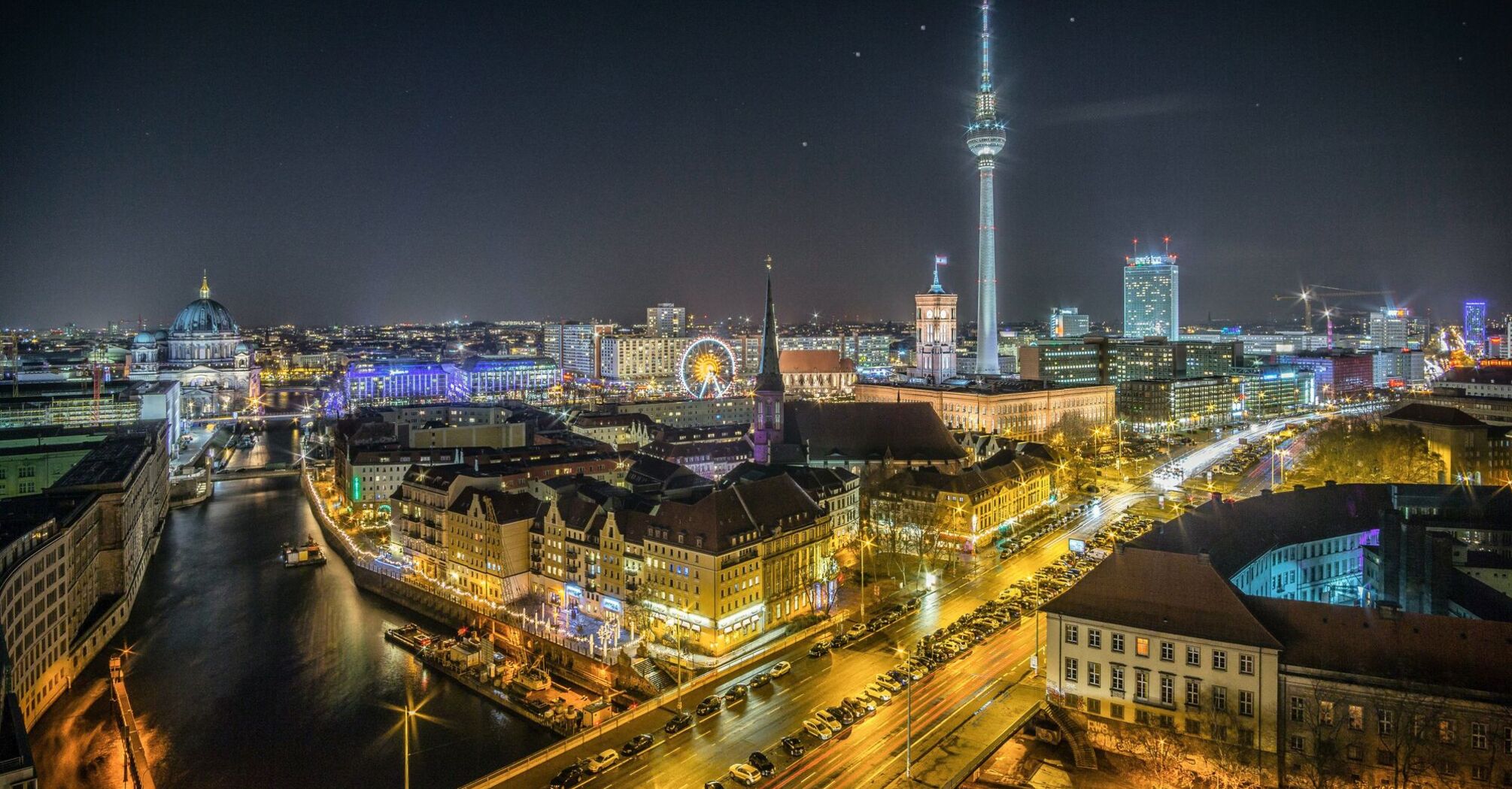 A night-time panoramic view of Berlin with the illuminated TV Tower in the backdrop, adjacent buildings, the Spree river, and busy streets with streaks of vehicle lights