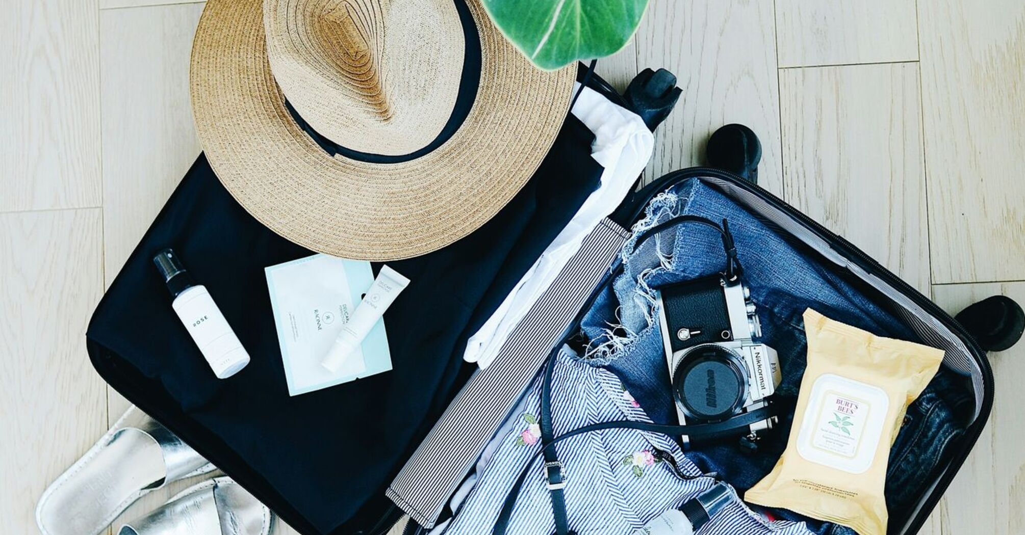 Open suitcase with clothing, camera, shoes, and toiletries, next to a wide-brimmed hat and a plant on a wooden floor