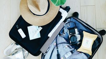 Open suitcase with clothing, camera, shoes, and toiletries, next to a wide-brimmed hat and a plant on a wooden floor