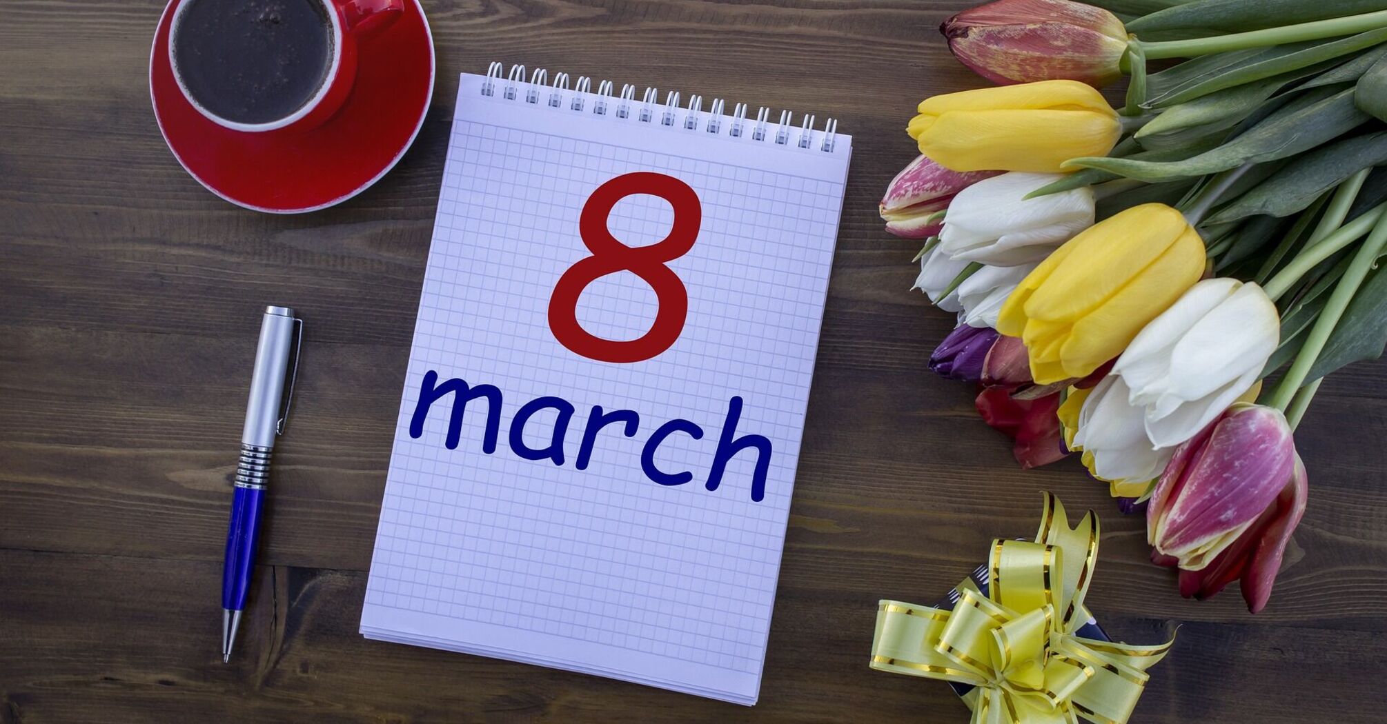 A notebook with "8 March" written on it, a red cup of coffee, a pen, and a bouquet of colorful tulips on a wooden background