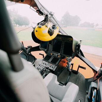 Interior view of a helicopter cockpit and instrument panels, with a pilot's yellow helmet hanging on the left 