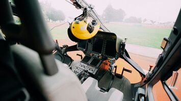 Interior view of a helicopter cockpit and instrument panels, with a pilot's yellow helmet hanging on the left 