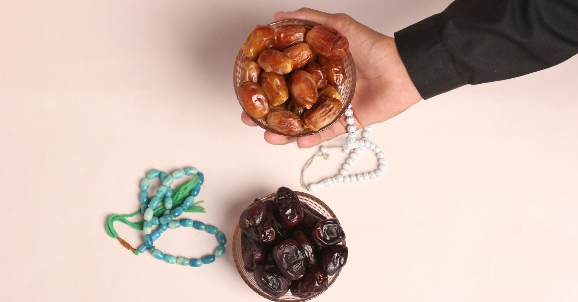 A hand holding a bowl of dates with prayer beads lying nearby on a plain surface 