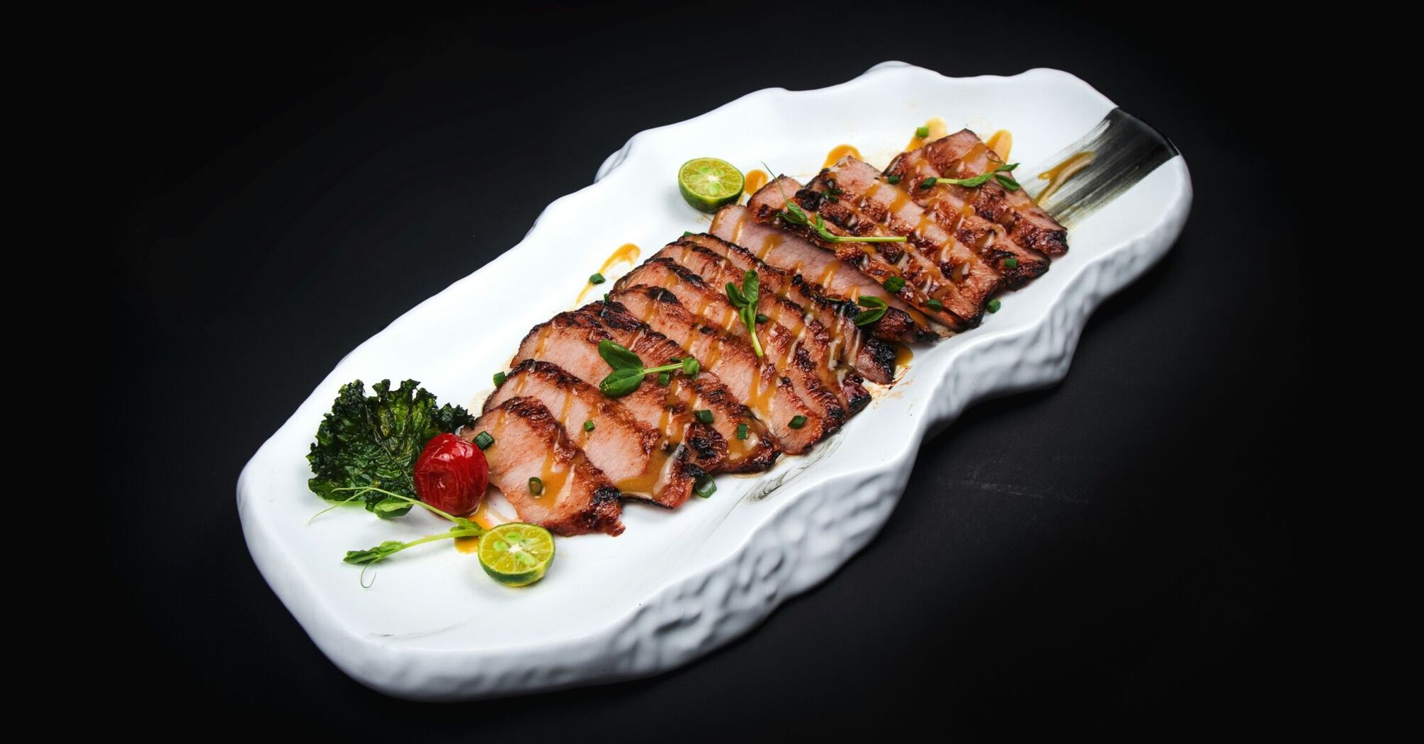 Sliced grilled pork on a white ceramic platter garnished with cherry tomatoes, kale, and lime 