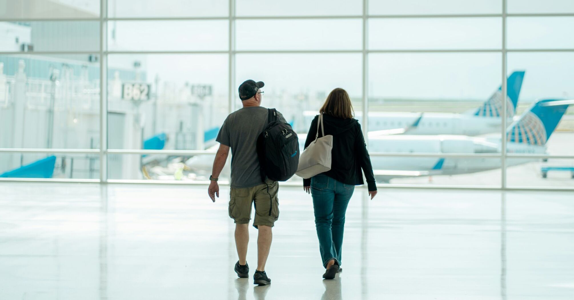 A man and a woman walking hand in hand in an airport terminal, with a clear view of parked airplanes through the large glass windows in the background