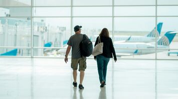 A man and a woman walking hand in hand in an airport terminal, with a clear view of parked airplanes through the large glass windows in the background