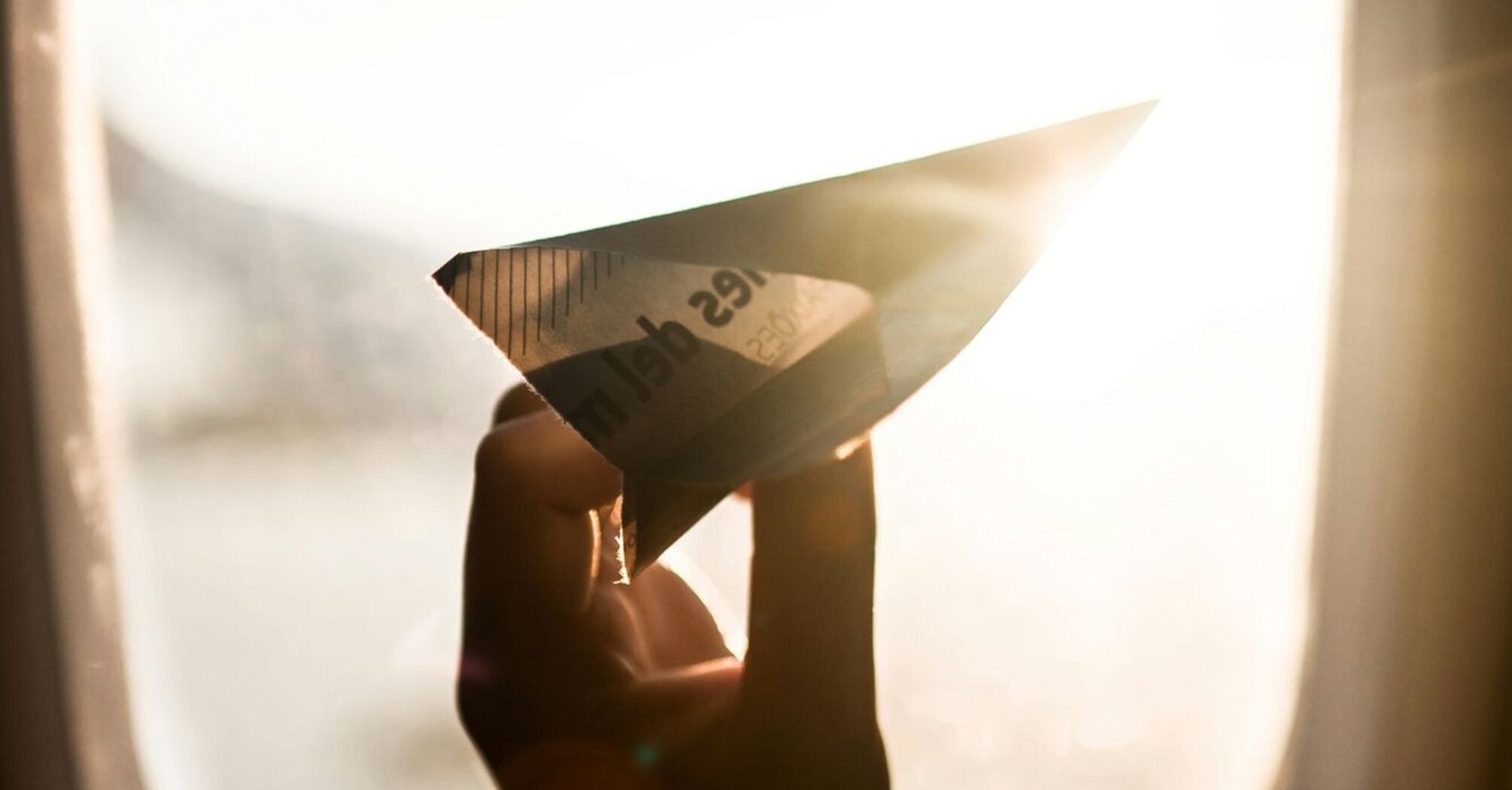 A hand holding a paper airplane against an airplane window with sunlight streaming