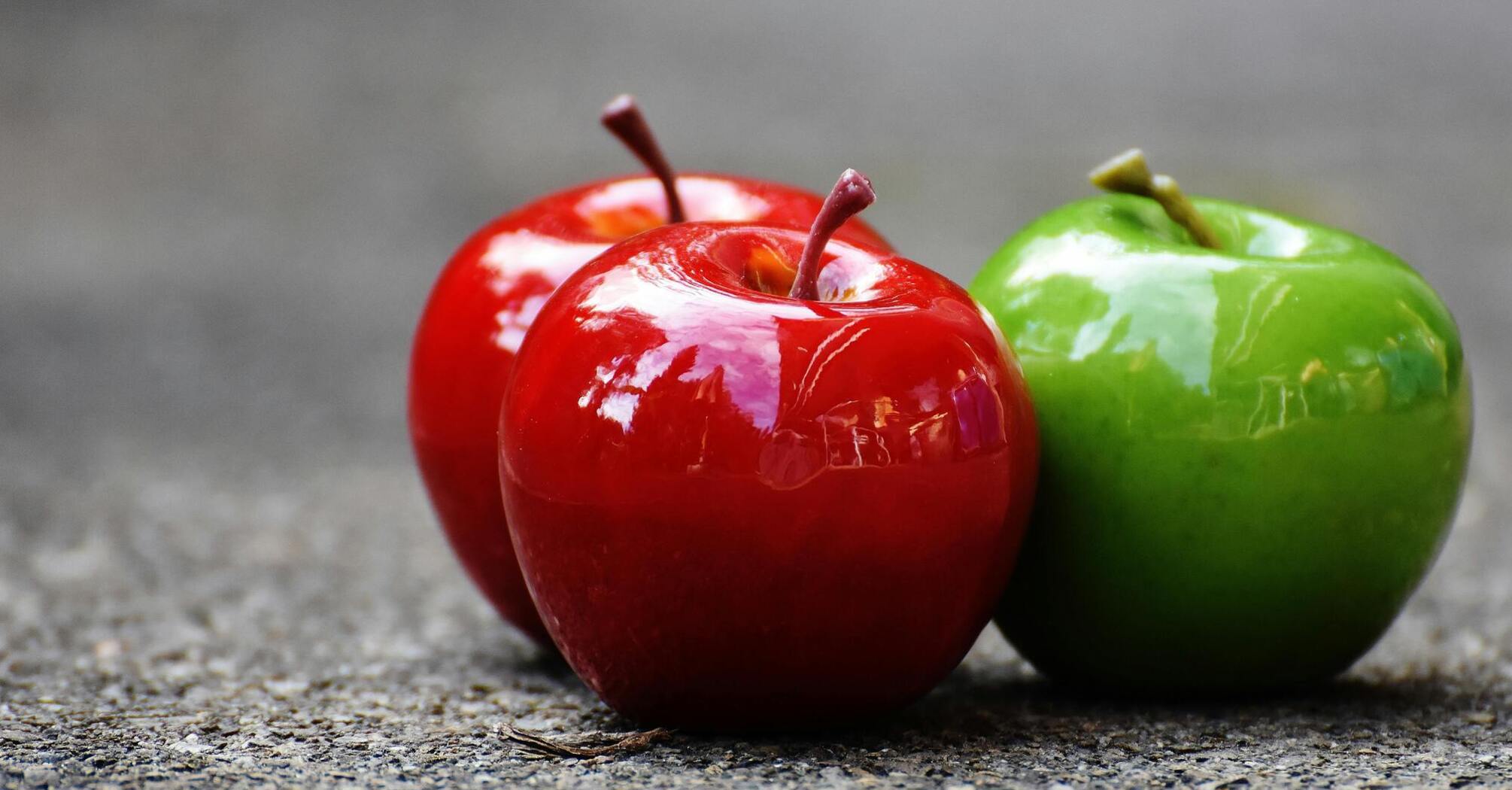 Three glossy apples on a concrete surface, with two red apples and one green apple