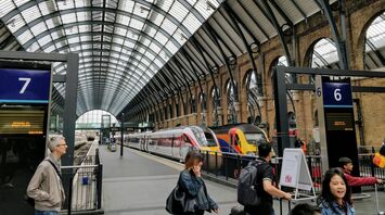 Interior of a busy train station with passengers, arched windows, and trains at the platforms under a curved glass roof 