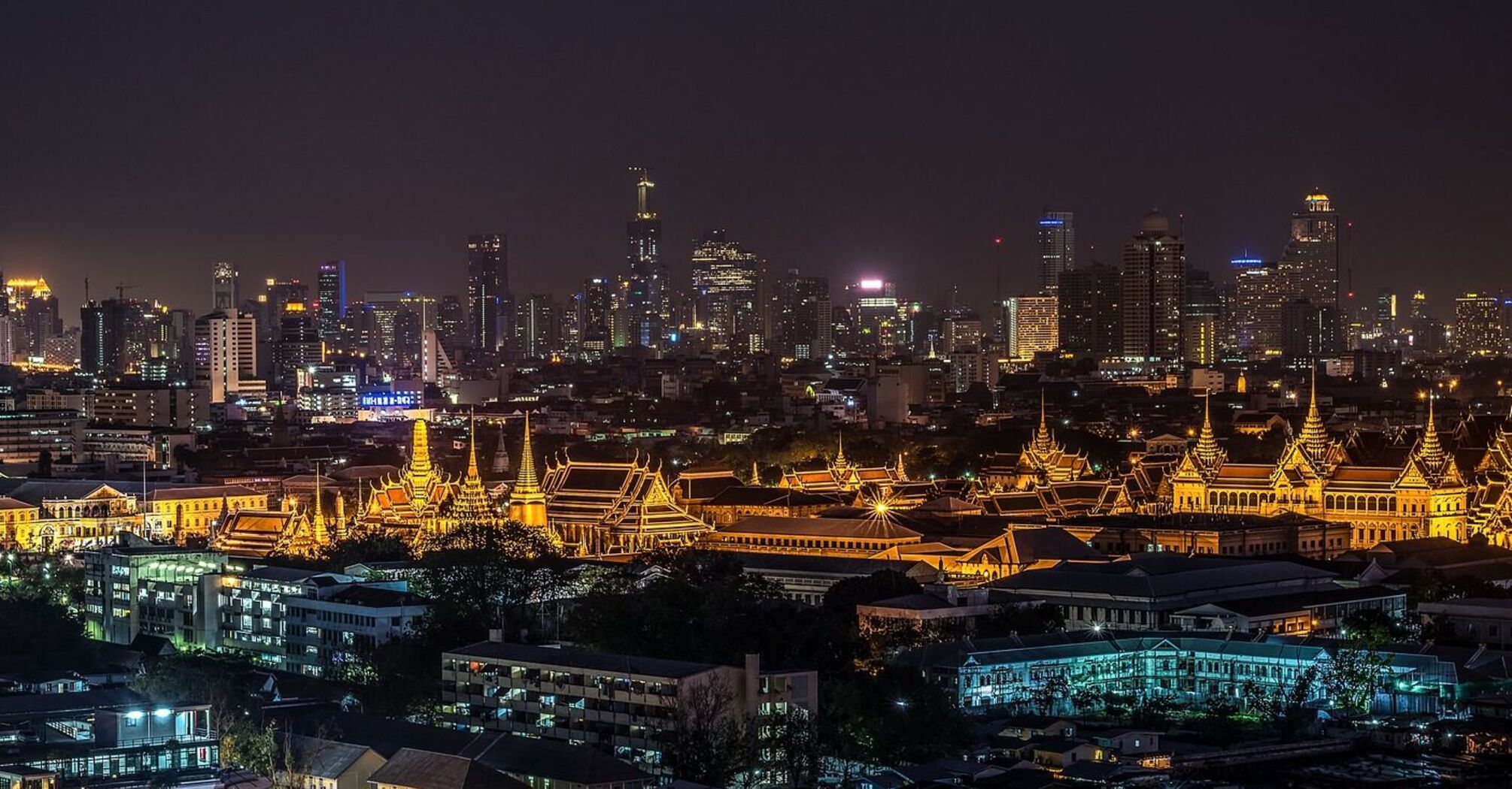 A panoramic night view of Bangkok's cityscape with illuminated Grand Palace and surrounding modern skyscrapers