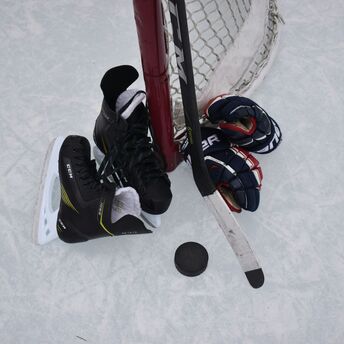 Hockey equipment including a pair of skates, gloves, and a stick resting against a goalpost on an ice rink, with a hockey puck on the ice