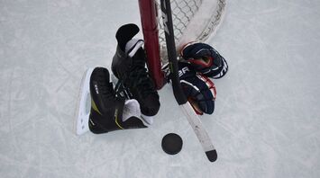 Hockey equipment including a pair of skates, gloves, and a stick resting against a goalpost on an ice rink, with a hockey puck on the ice