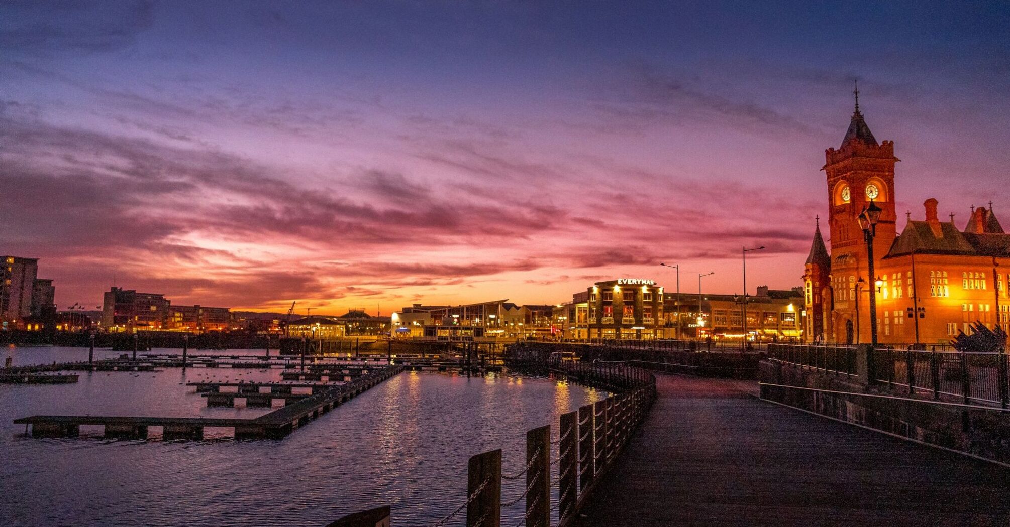 A vibrant sunset over Cardiff Bay with silhouettes of buildings and the Pierhead Building clock tower, reflecting in the water