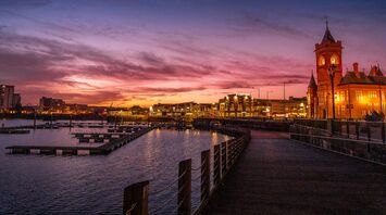 A vibrant sunset over Cardiff Bay with silhouettes of buildings and the Pierhead Building clock tower, reflecting in the water