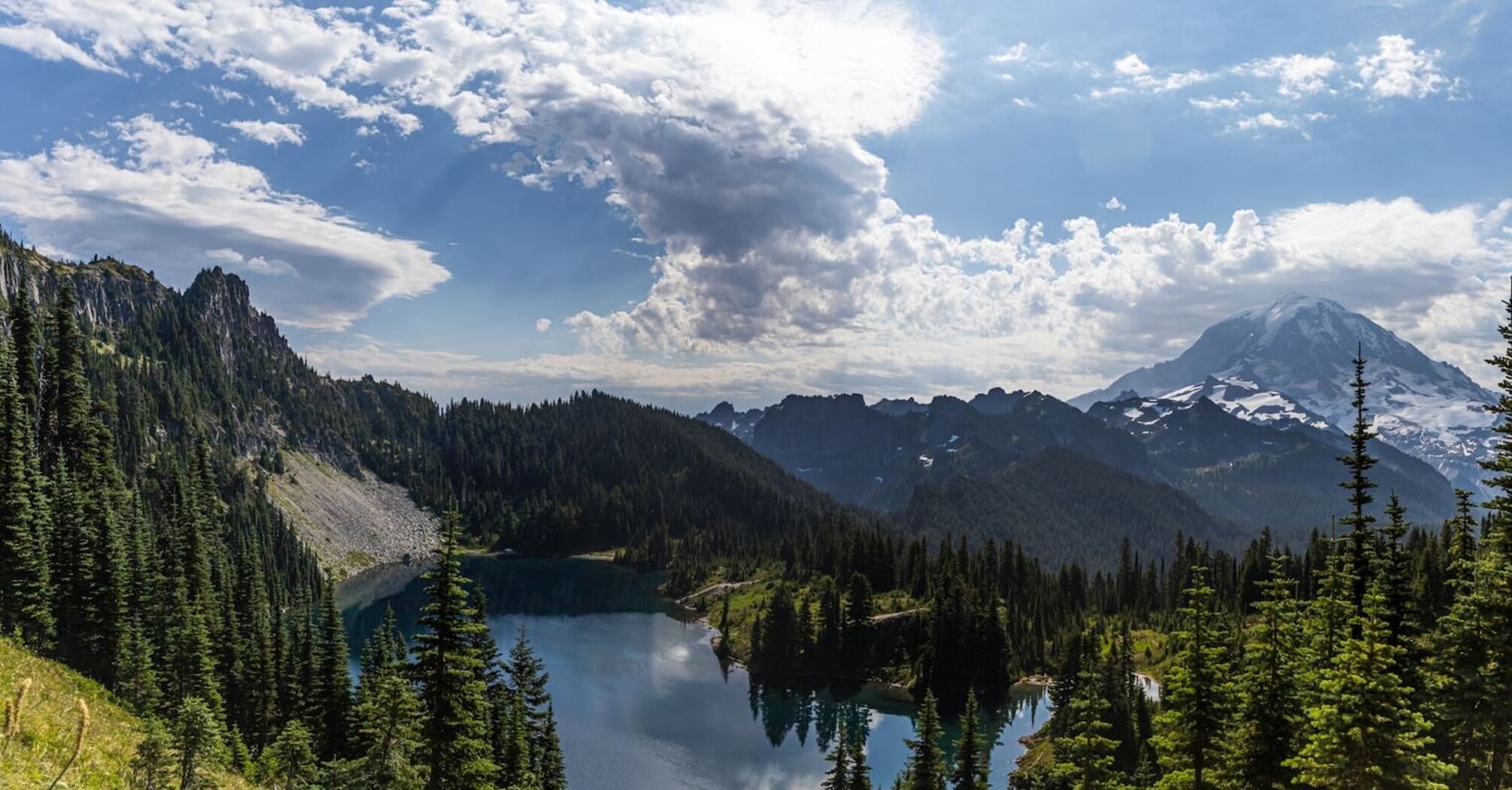 A panoramic view of a mountain with a lake, forests, and clouds