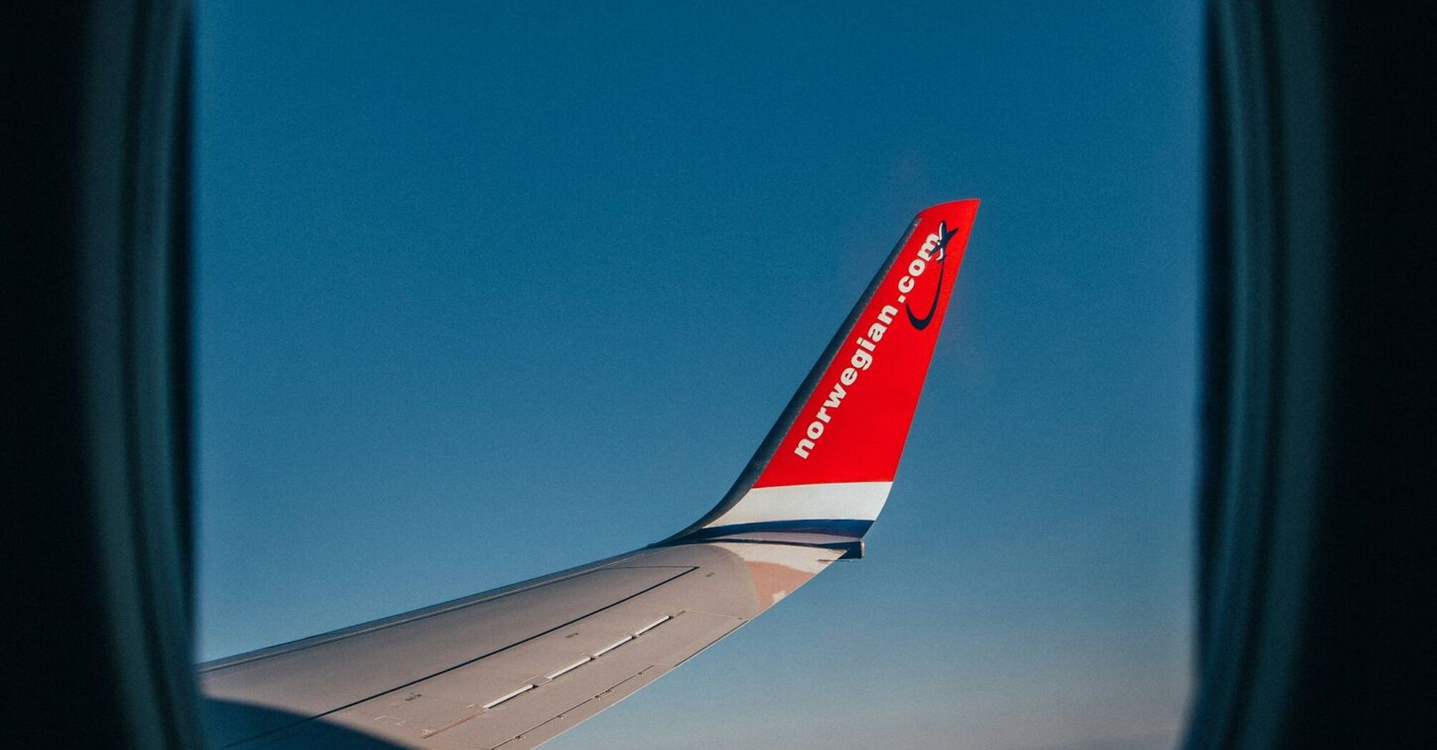 View from an airplane window showing a wing with the Norwegian Air logo against a clear blue sky