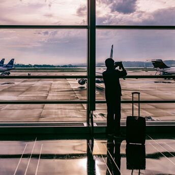 A silhouette of a person taking a photo at an airport terminal, with planes visible through large windows and a suitcase beside them
