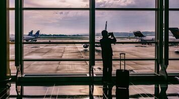 A silhouette of a person taking a photo at an airport terminal, with planes visible through large windows and a suitcase beside them