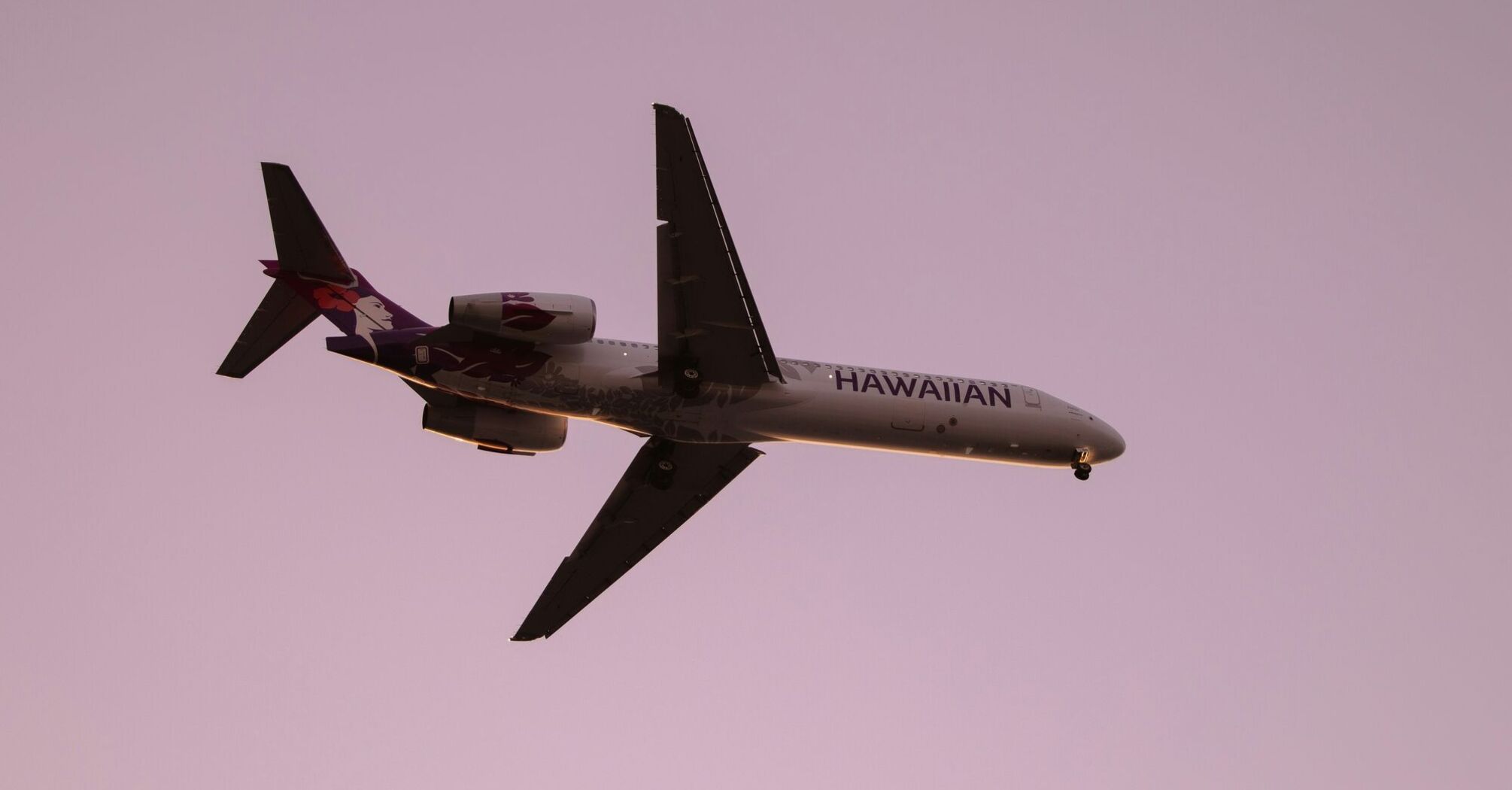 A Hawaiian Airlines aircraft in flight against a pink sky backdrop