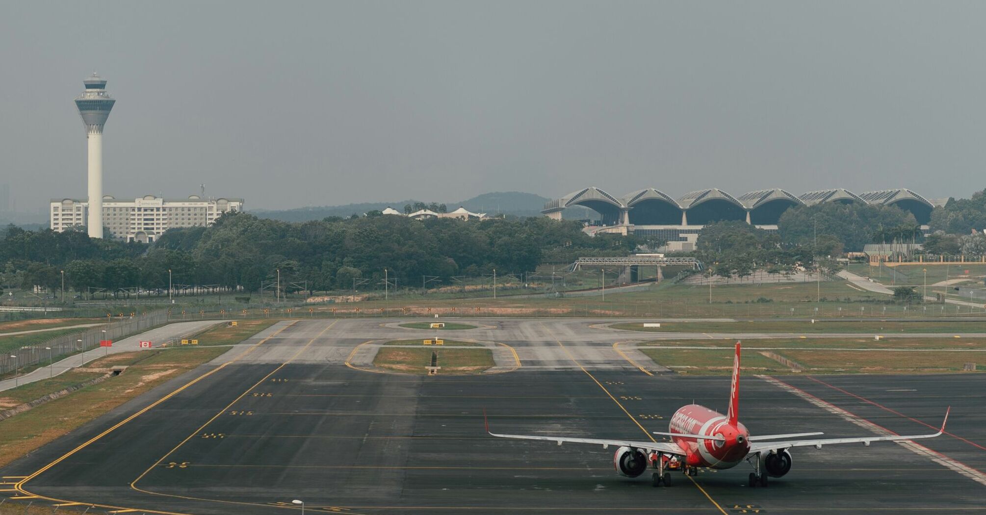 A red AirAsia airplane on a runway, with an airport control tower and terminal buildings in the background, set against a hazy sky 