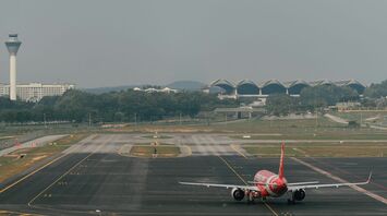 A red AirAsia airplane on a runway, with an airport control tower and terminal buildings in the background, set against a hazy sky