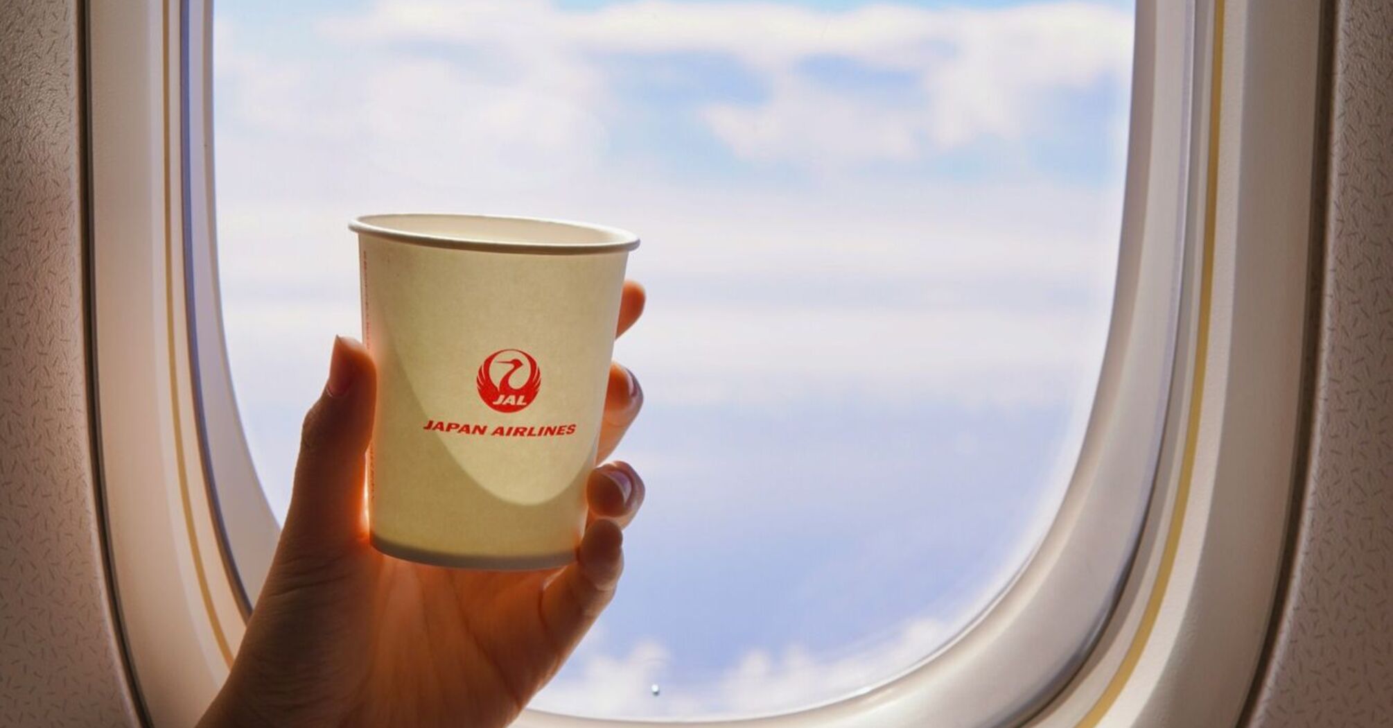 A person holding a Japan Airlines (JAL) cup near an airplane window, with a view of the sky and clouds outside