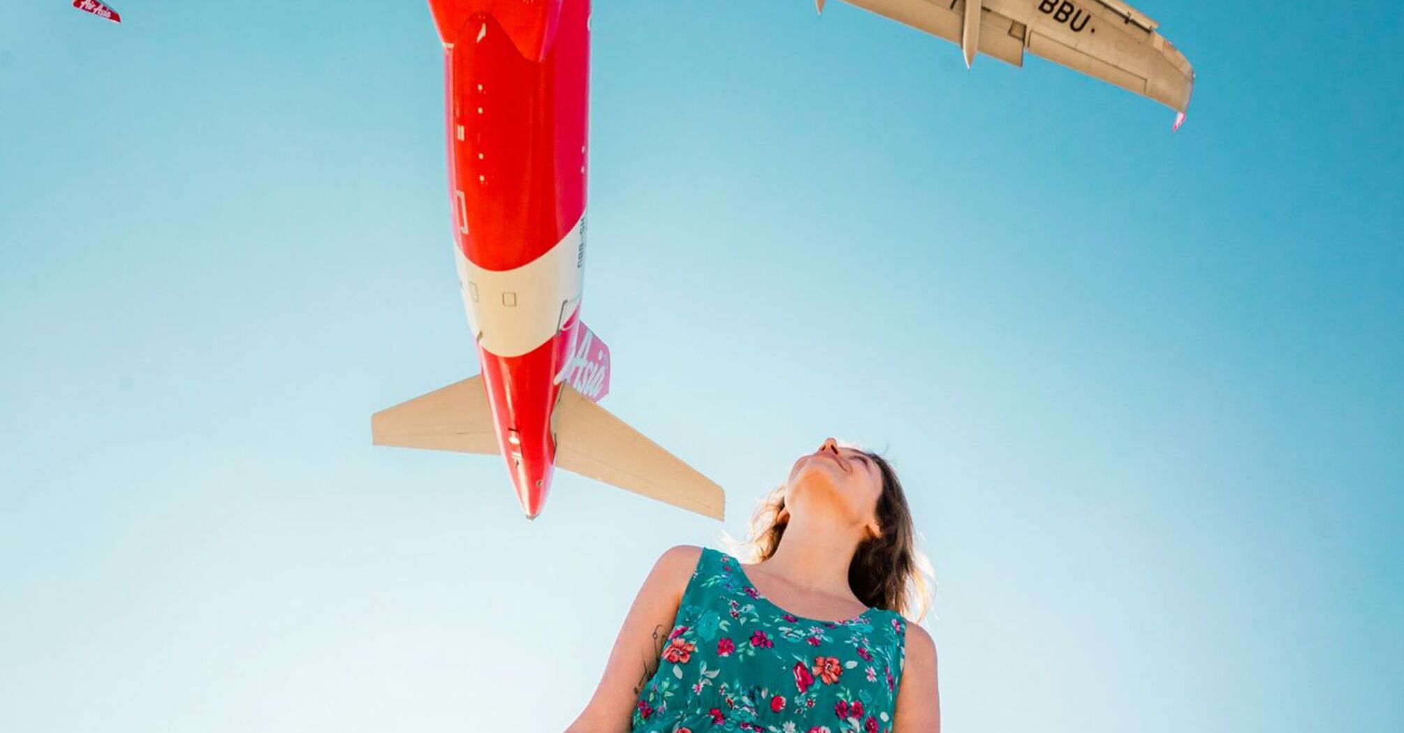 A person in a floral dress stands beneath an AirAsia plane flying overhead against a clear blue sky