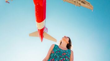 A person in a floral dress stands beneath an AirAsia plane flying overhead against a clear blue sky