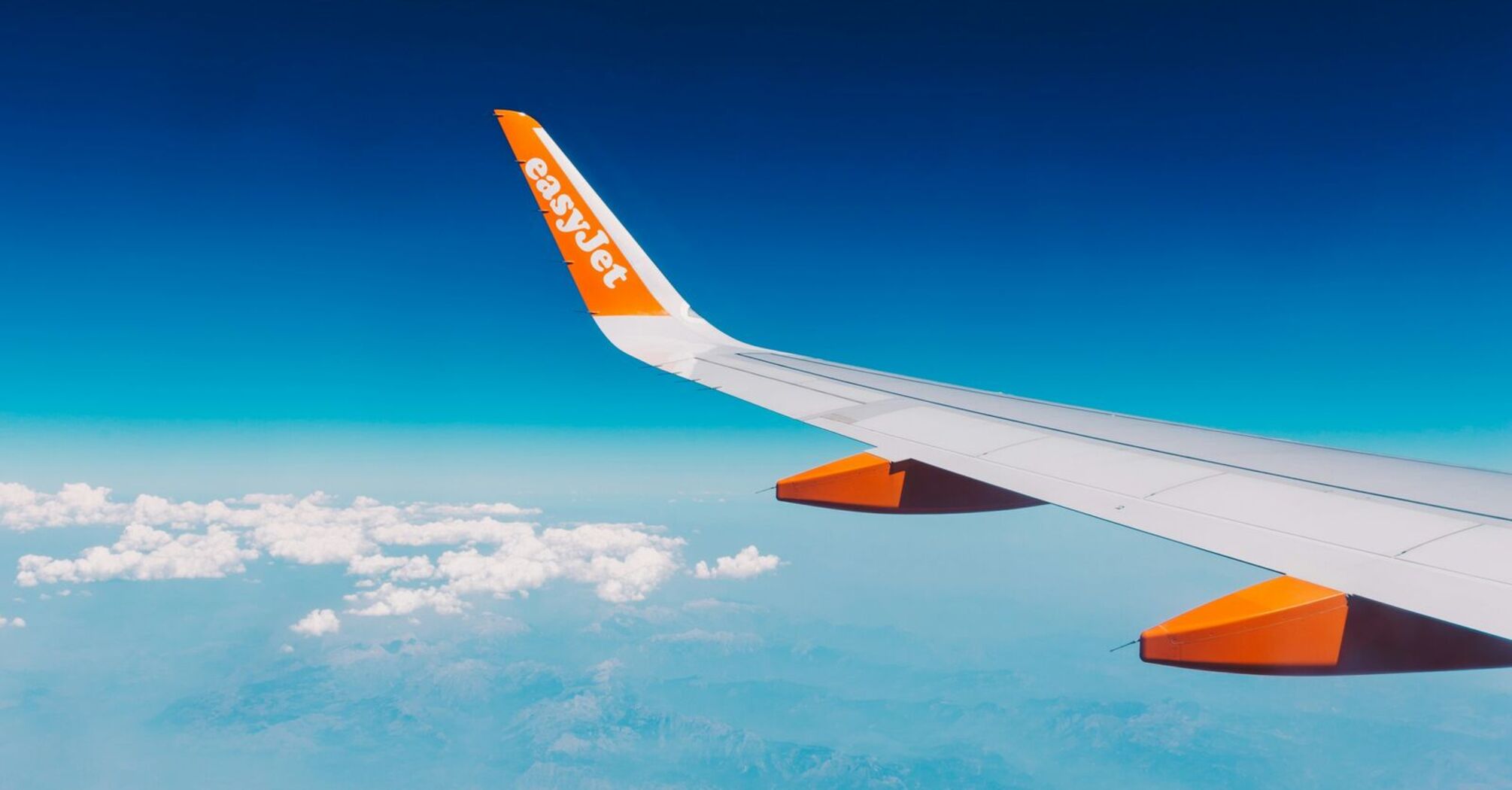 An airplane wing with the easyJet logo flying over a landscape with clouds and blue sky 