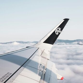 An airplane wing with an Air New Zealand logo, flying above a sea of clouds with mountains in the distance