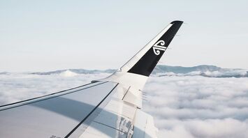 An airplane wing with an Air New Zealand logo, flying above a sea of clouds with mountains in the distance