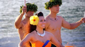 A Hawaiian dancer with a yellow hibiscus flower in her hair, accompanied by two men with leaf crowns, stands near the water