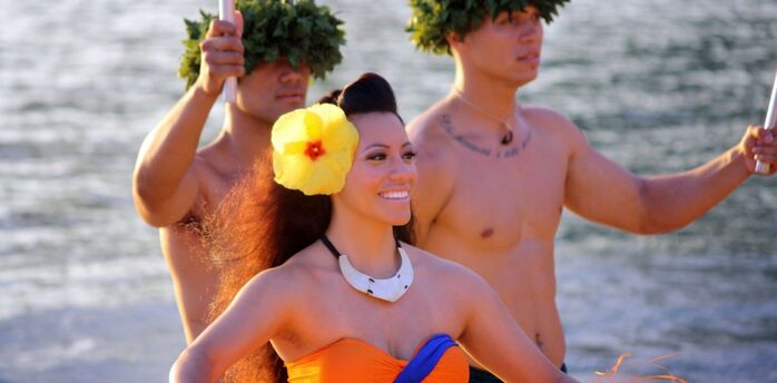 A Hawaiian dancer with a yellow hibiscus flower in her hair, accompanied by two men with leaf crowns, stands near the water