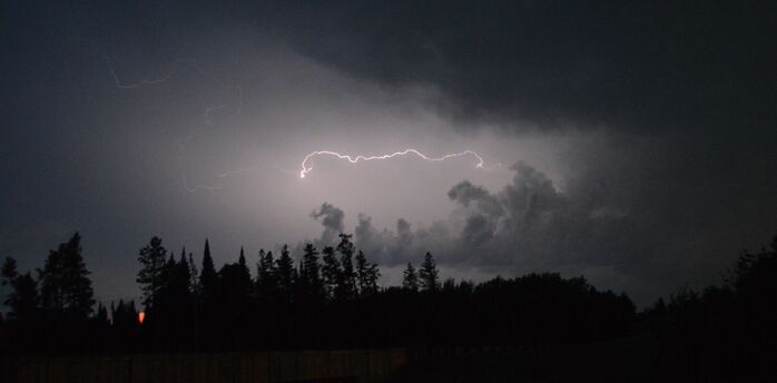 A dark stormy sky illuminated by a streak of lightning, with silhouettes of trees in the foreground