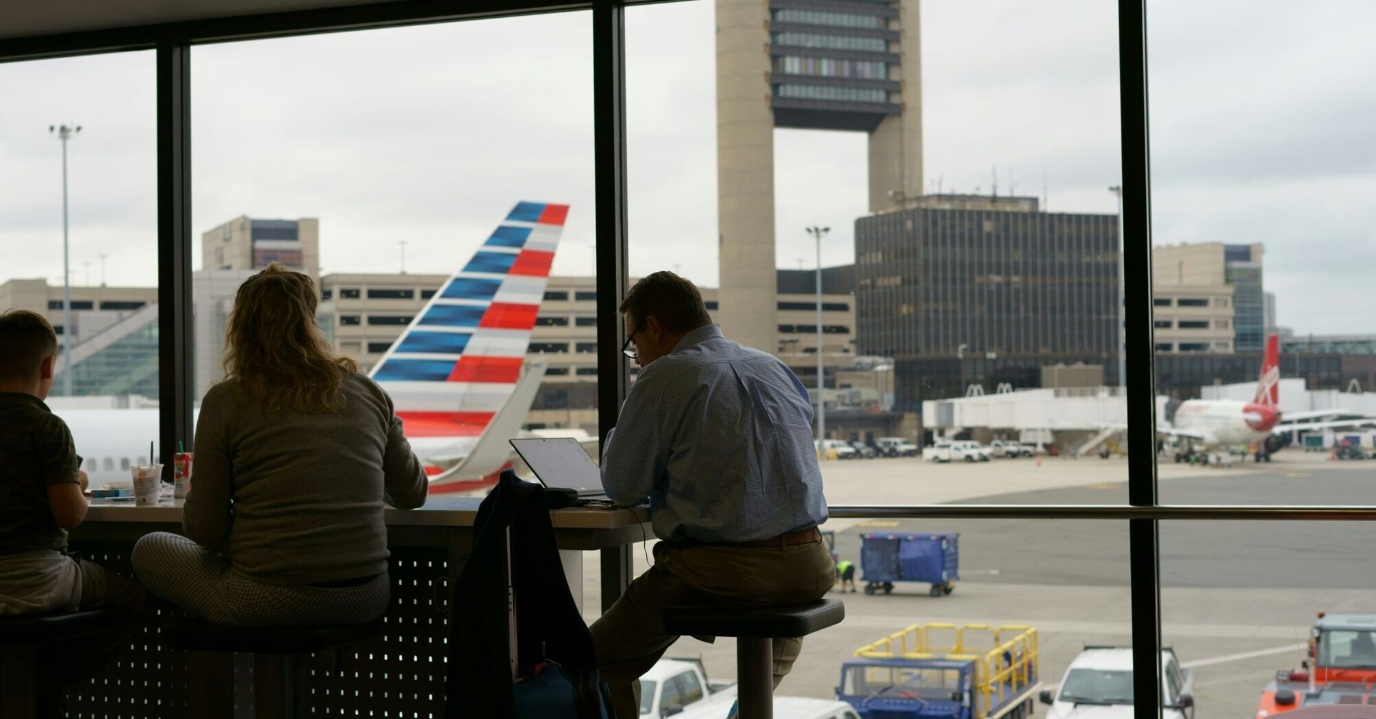 Passengers seated at a bar table inside Boston Logan International Airport with a view of the airfield and aircraft