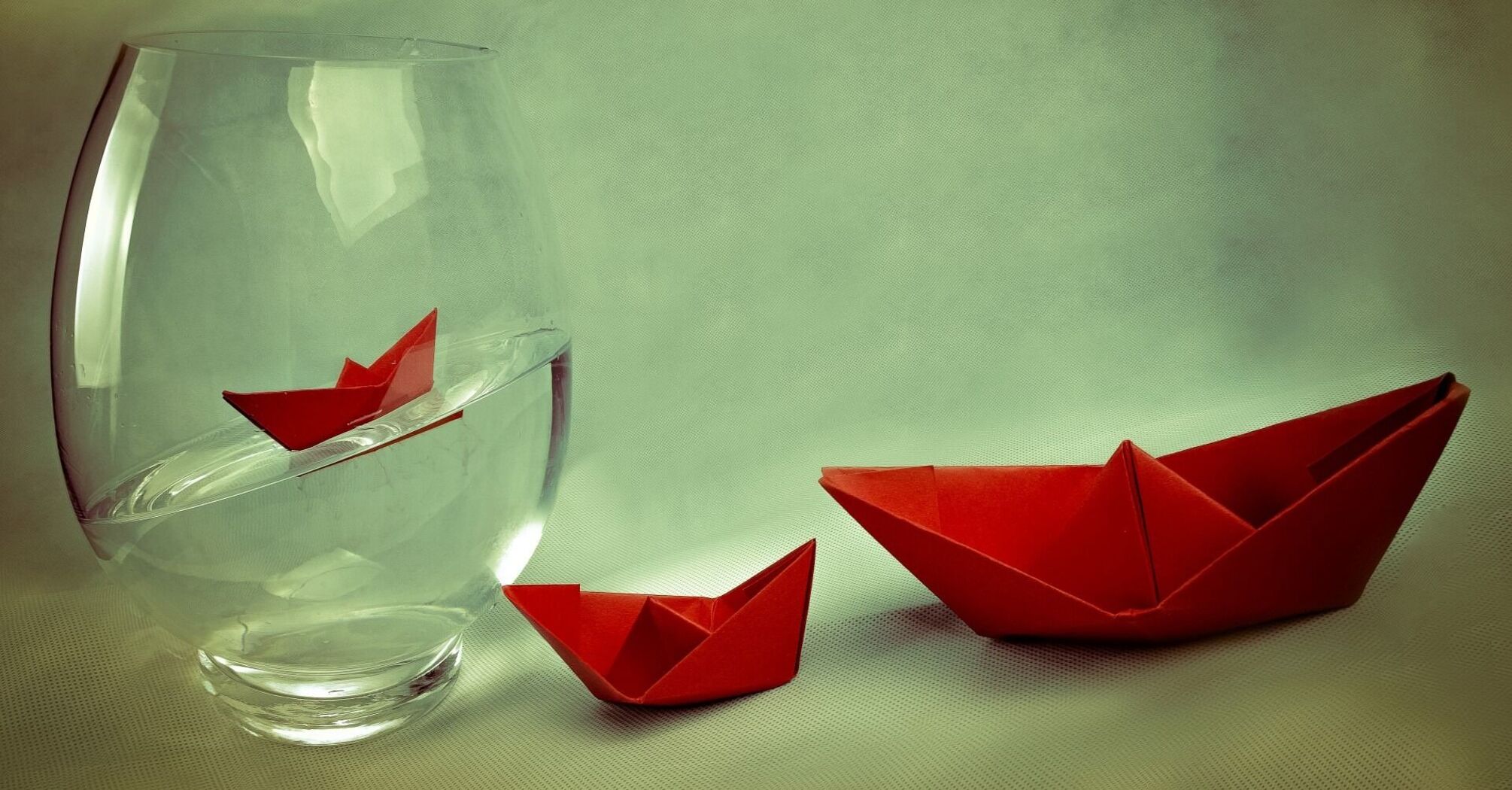 A red paper boat inside a large glass vase half-filled with water, with another red paper boat lying outside the vase on a beige surface