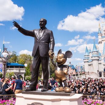 Statue of Walt Disney holding hands with Mickey Mouse in front of Cinderella Castle at a crowded Disney theme park