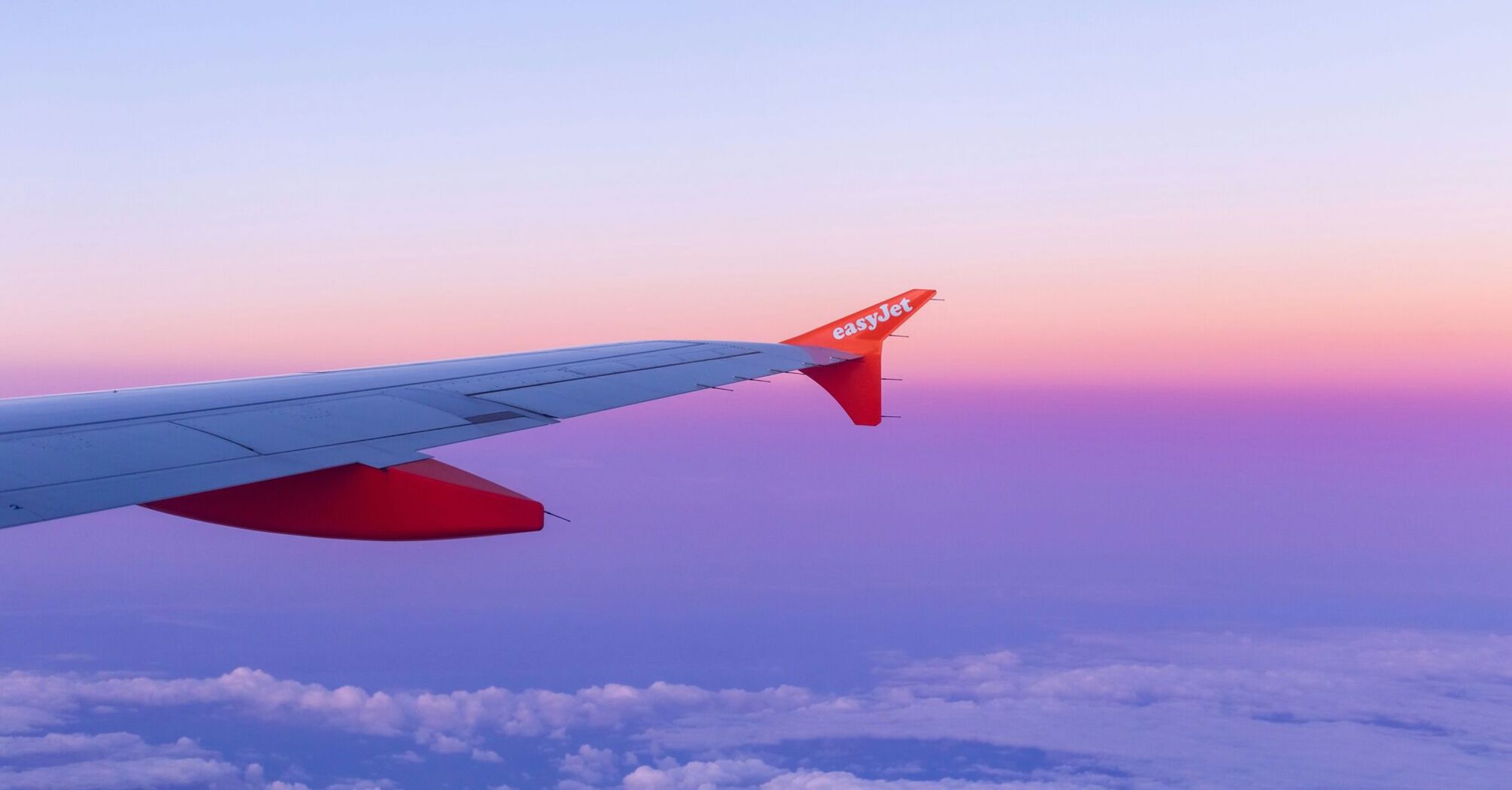 Wing of an easyJet airplane against a purple and pink sky at sunset, flying above the clouds
