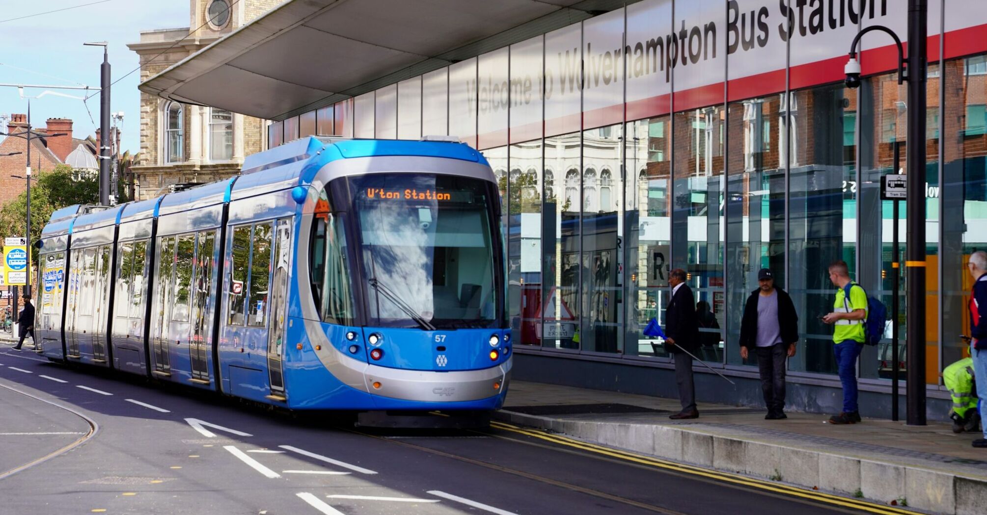 A modern blue tram in front of the Wolverhampton Bus Station, with pedestrians and workers nearby