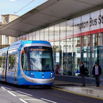 A modern blue tram in front of the Wolverhampton Bus Station, with pedestrians and workers nearby