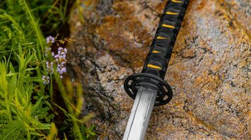 Sword with a black and yellow handle resting on a rocky surface surrounded by green plants