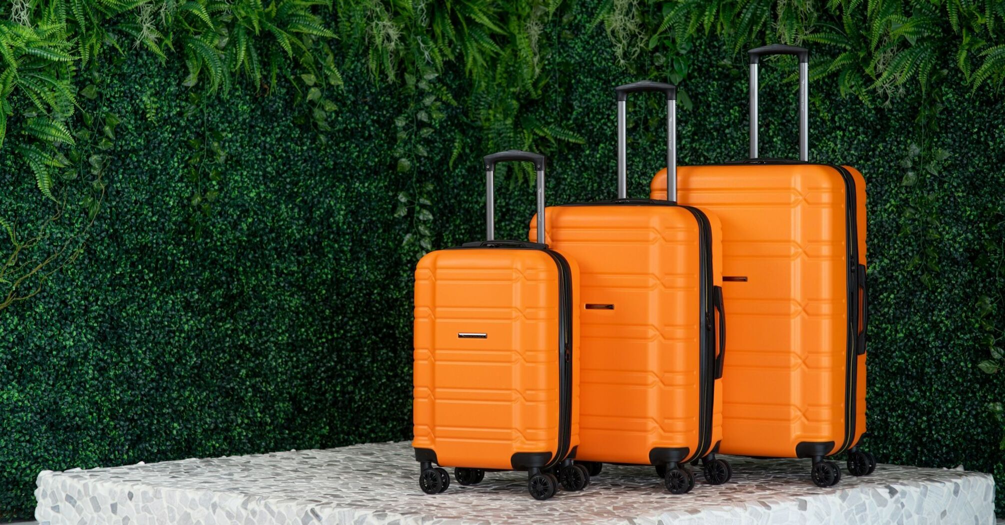 Three bright orange suitcases of varying sizes standing against a lush green plant-covered wall 