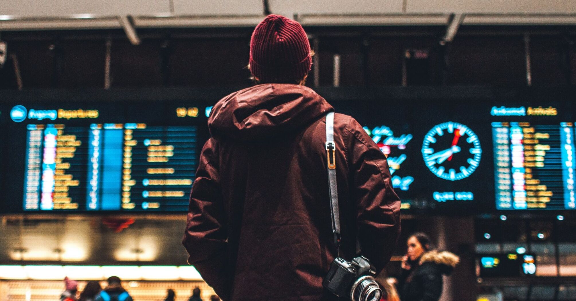 A traveler in a maroon jacket and beanie stands in front of an airport departure board