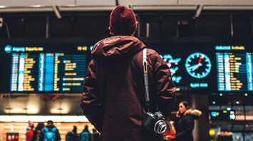 A traveler in a maroon jacket and beanie stands in front of an airport departure board
