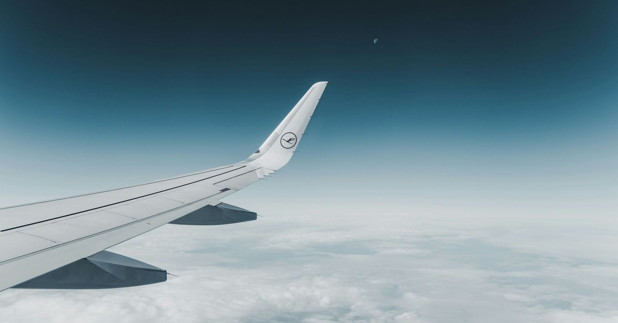 View from the wing of an airplane flying high above the clouds with the Lufthansa logo visible on the wingtip, clear skies and the moon in the distance