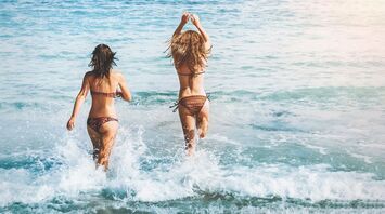 Two women running into the ocean waves at a beach, seen from behind, during a sunny day
