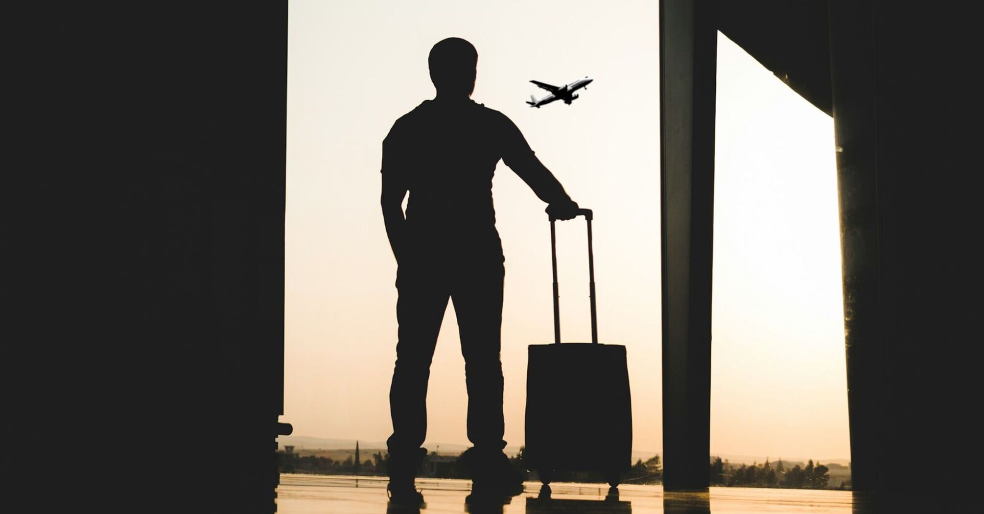 Silhouette of a man with a suitcase standing in an airport terminal, watching a plane take off during sunset