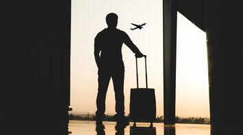 Silhouette of a man with a suitcase standing in an airport terminal, watching a plane take off during sunset